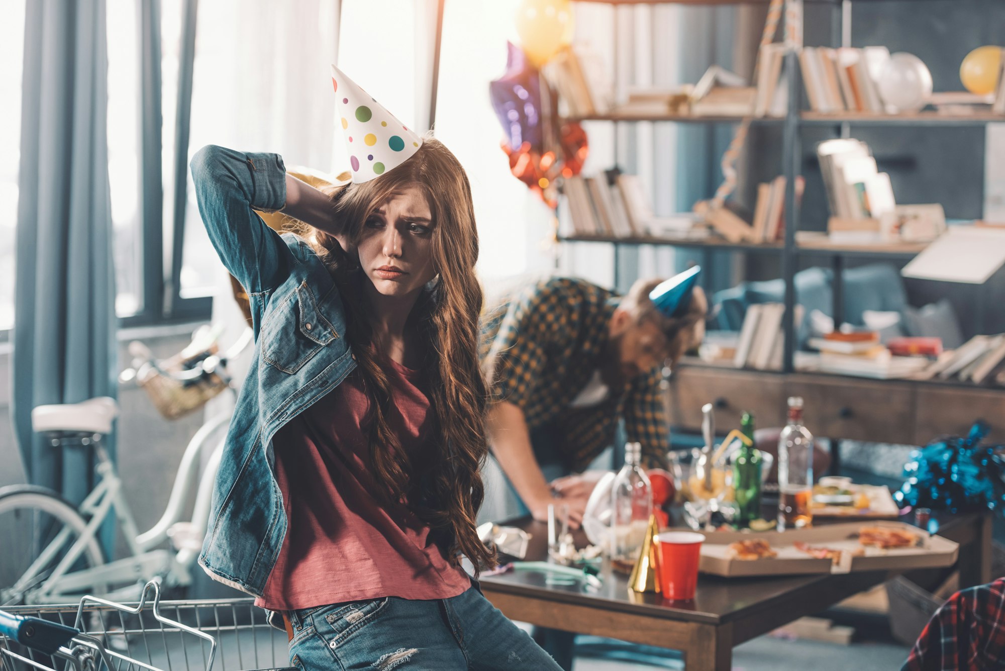 confused woman in birthday hat, man cleaning behind in messy room after party